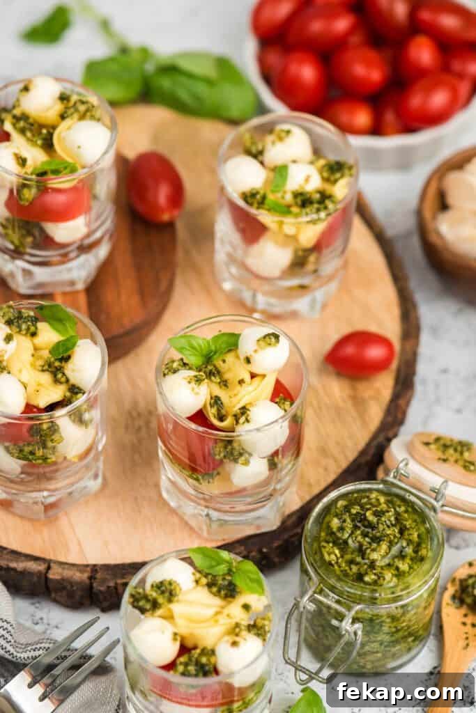 Tortellini Caprese salad cups on a wooden slab surrounded by fresh pesto.
