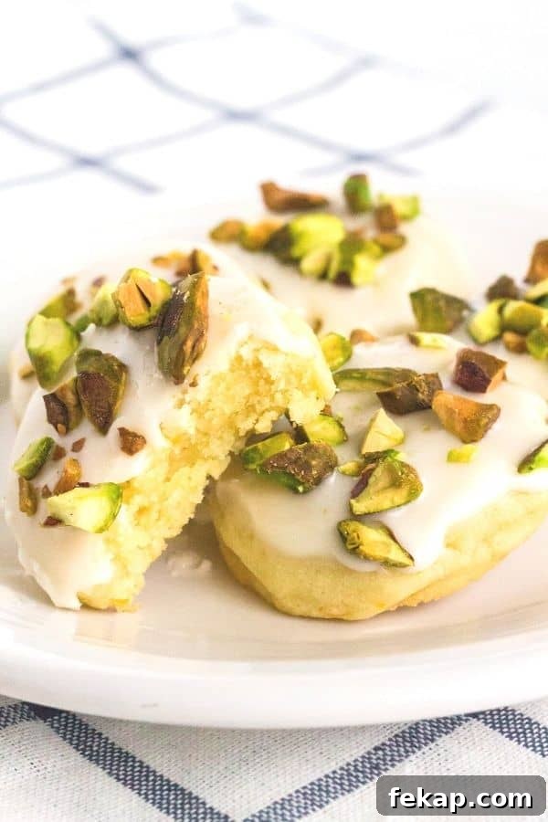 a few frosted lemon cookies topped with pistachios on a white plate, with a bite taken out of one cookie showing its tender interior