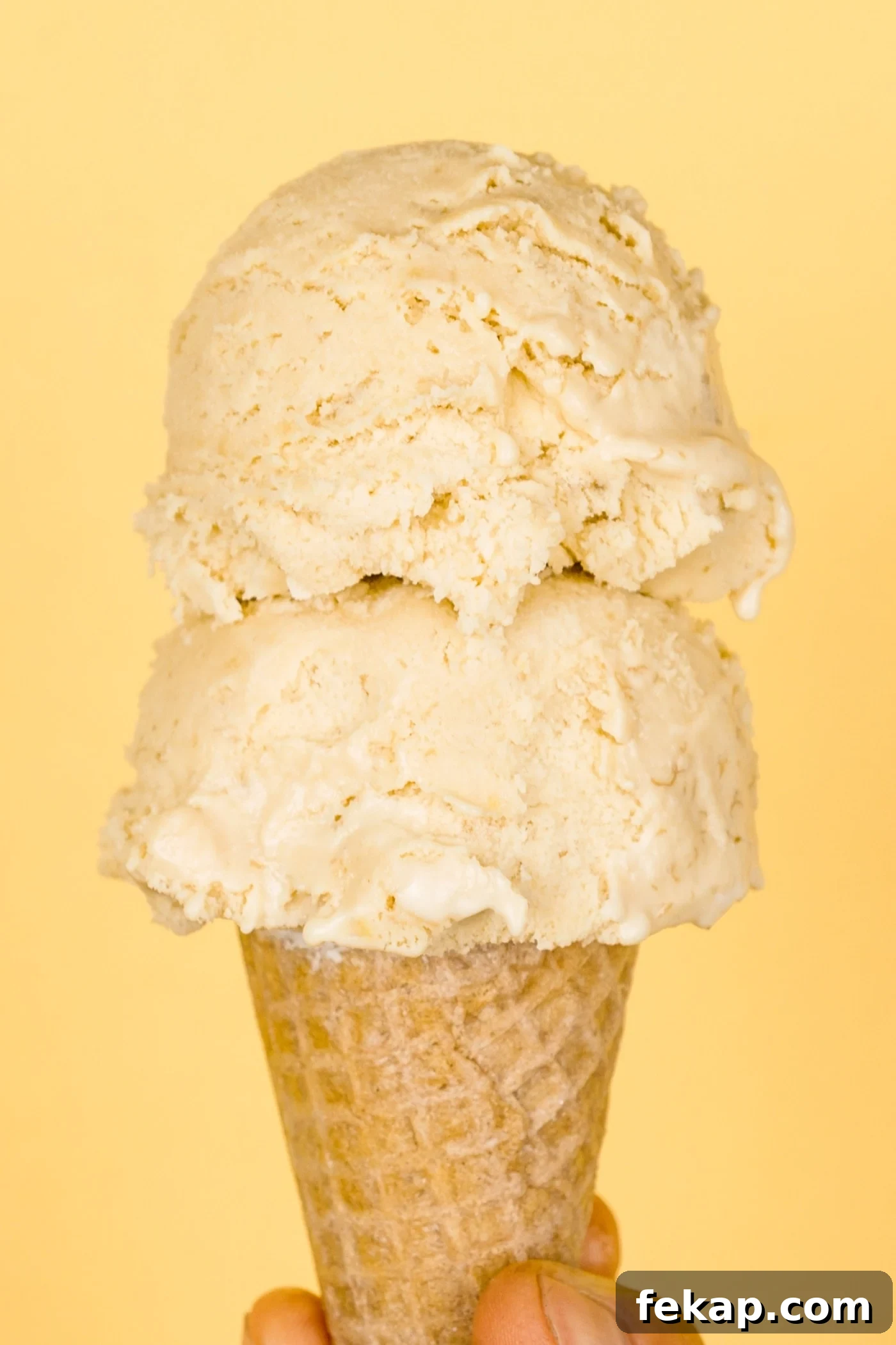 Homemade Banana Cream Pie Ice Cream Recipe 3 Two scoops of banana cream pie ice cream in ice cream cone held by a hand with light peach colored background