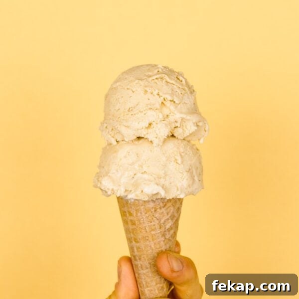 Two scoops of banana cream pie ice cream in ice cream cone held by a hand with light peach colored background