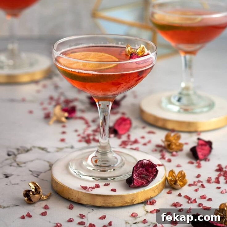 Strawberry gimlet garnished with a slice of lime, on a white and gold coaster with pink petals and rocks scattered at the base.