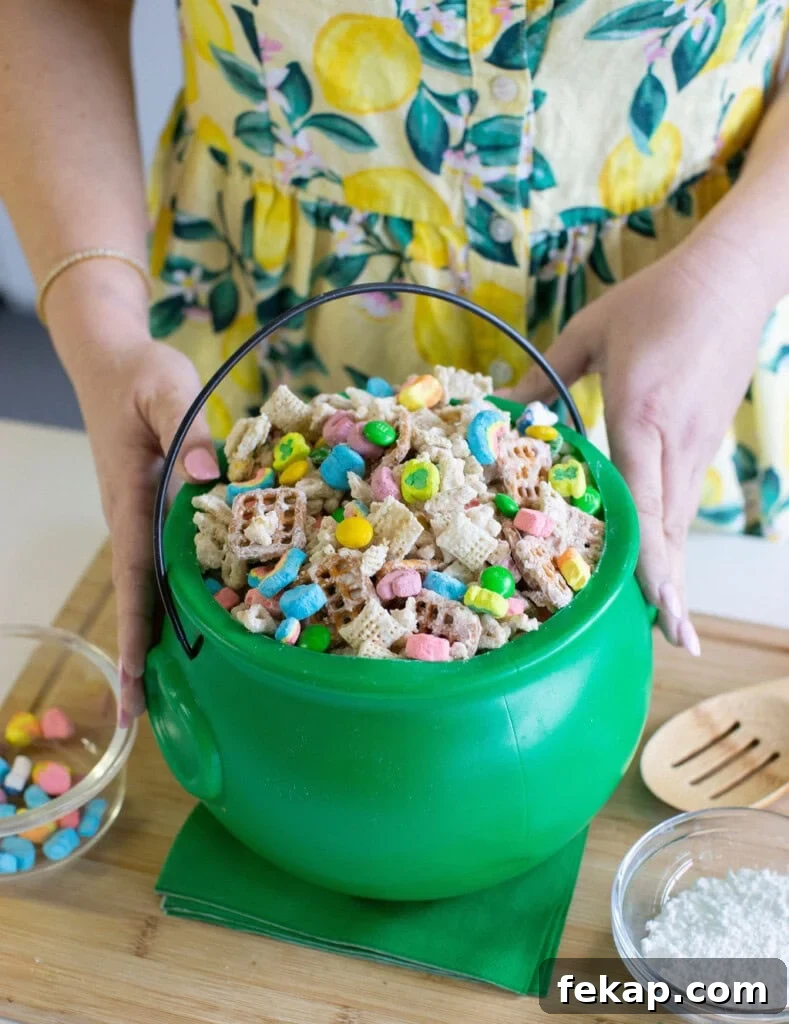Lucky Charms Puppy Chow: Leprechaun Bait Recipe 2 A person in a yellow, lemon-patterned dress holds a green plastic cauldron filled with Chex cereal and colorful marshmallows, with a small bowl of marshmallows and a bowl of powdered sugar nearby.