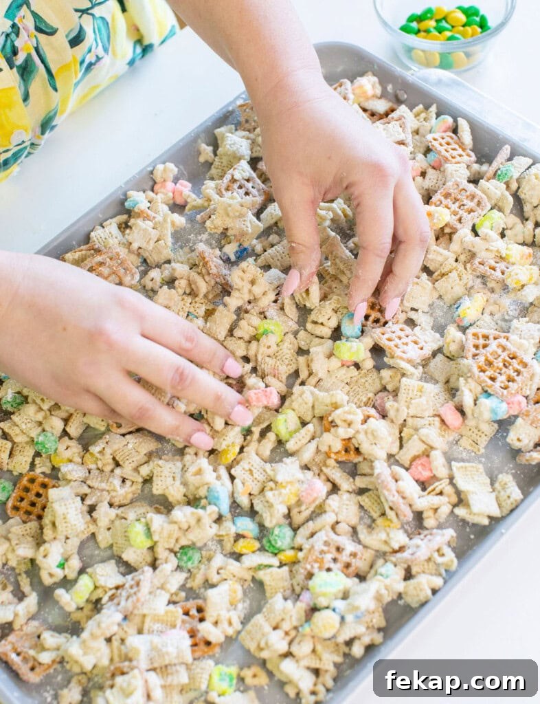 Lucky Charms Puppy Chow: Leprechaun Bait Recipe 4 A person arranges a cereal snack mix with colorful candies, pretzels, and assorted cereals on a baking sheet. A small bowl of additional candies sits nearby on a white surface.