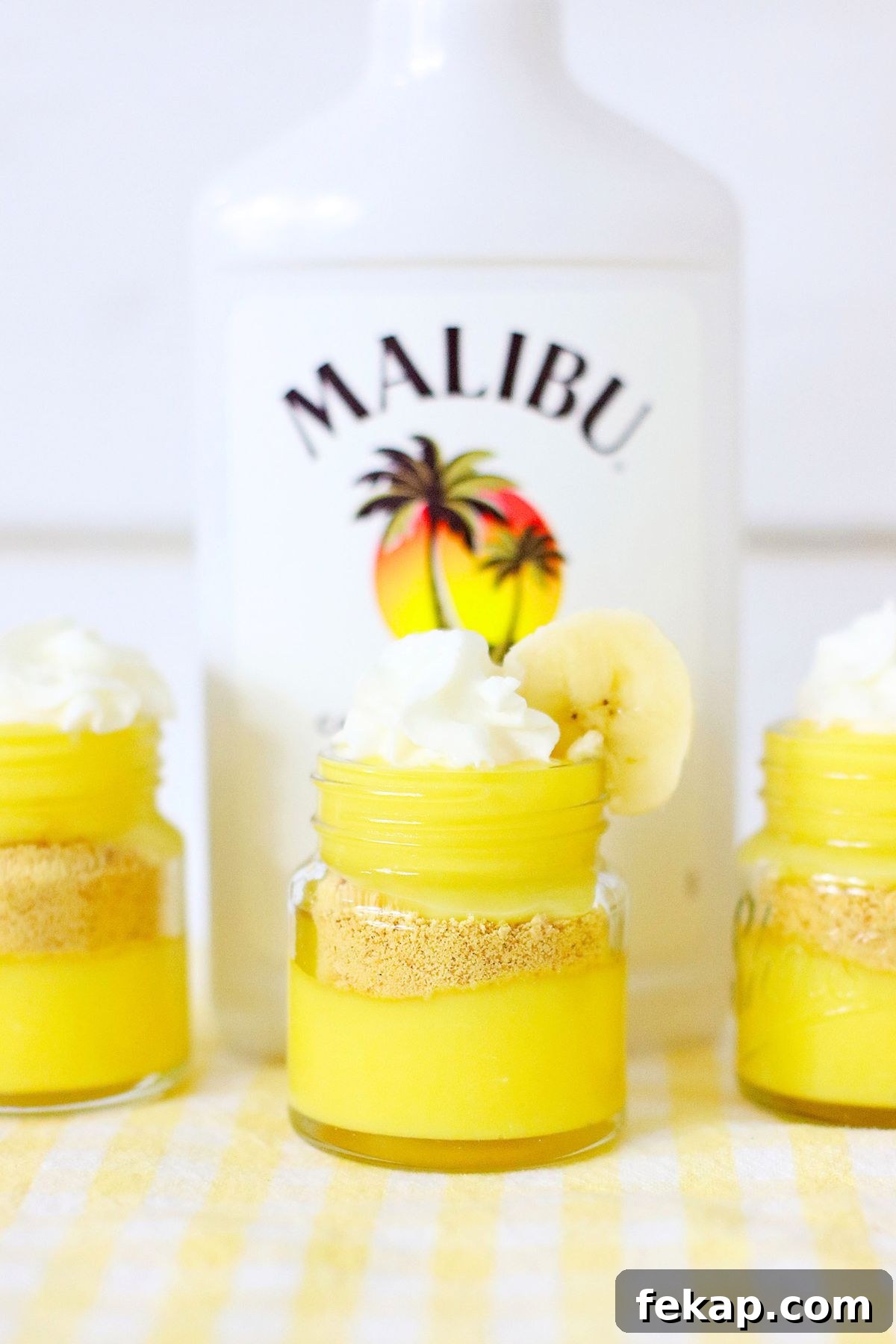 Creamy Banana Pudding Shots Recipe for Parties 6 Bright yellow pudding layers in mini jars, topped with whipped cream and garnished with a banana slice in front of a bottle of rum.