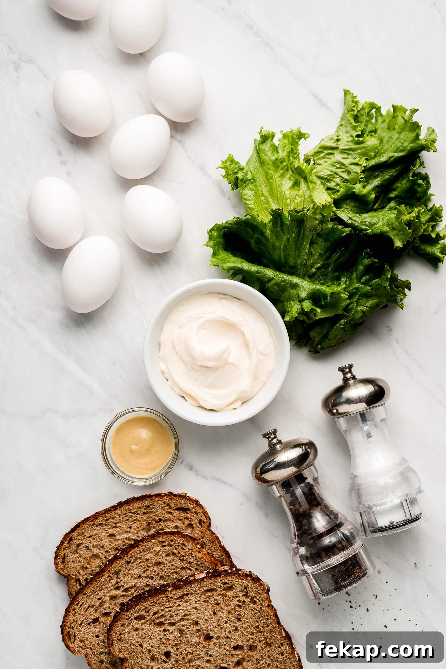 Ingredients on a marble surface- eggs, lettuce, mayo, mustard, bread, and salt & pepper.