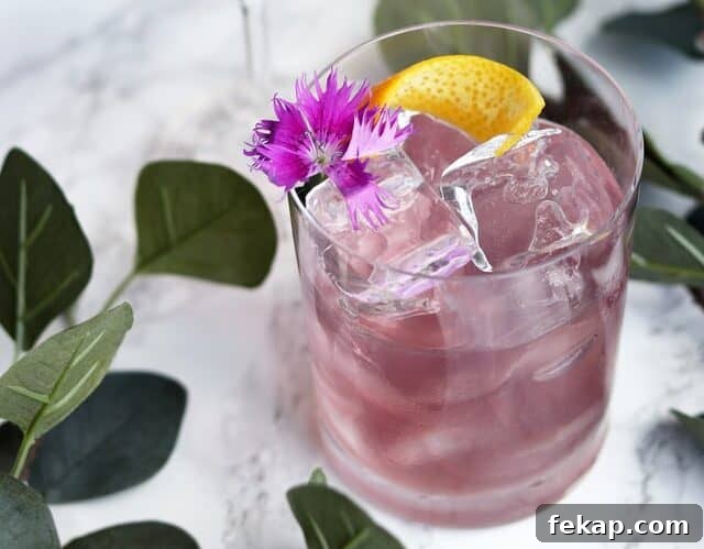 A purple cocktail garnished with a flower and lemon on a white marble table.