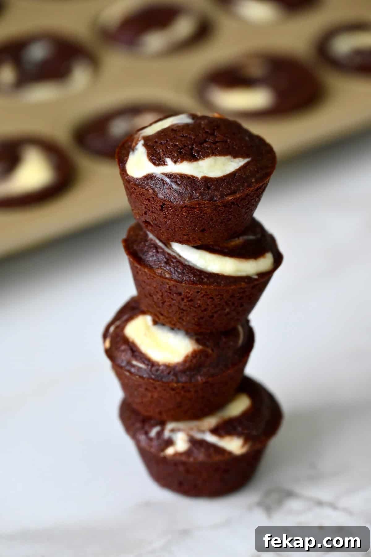 Fudgy Chocolate Brownies Recipe 12 Cheesecake brownie bites are stacked on a marble counter, with a mini muffin pan in the background.