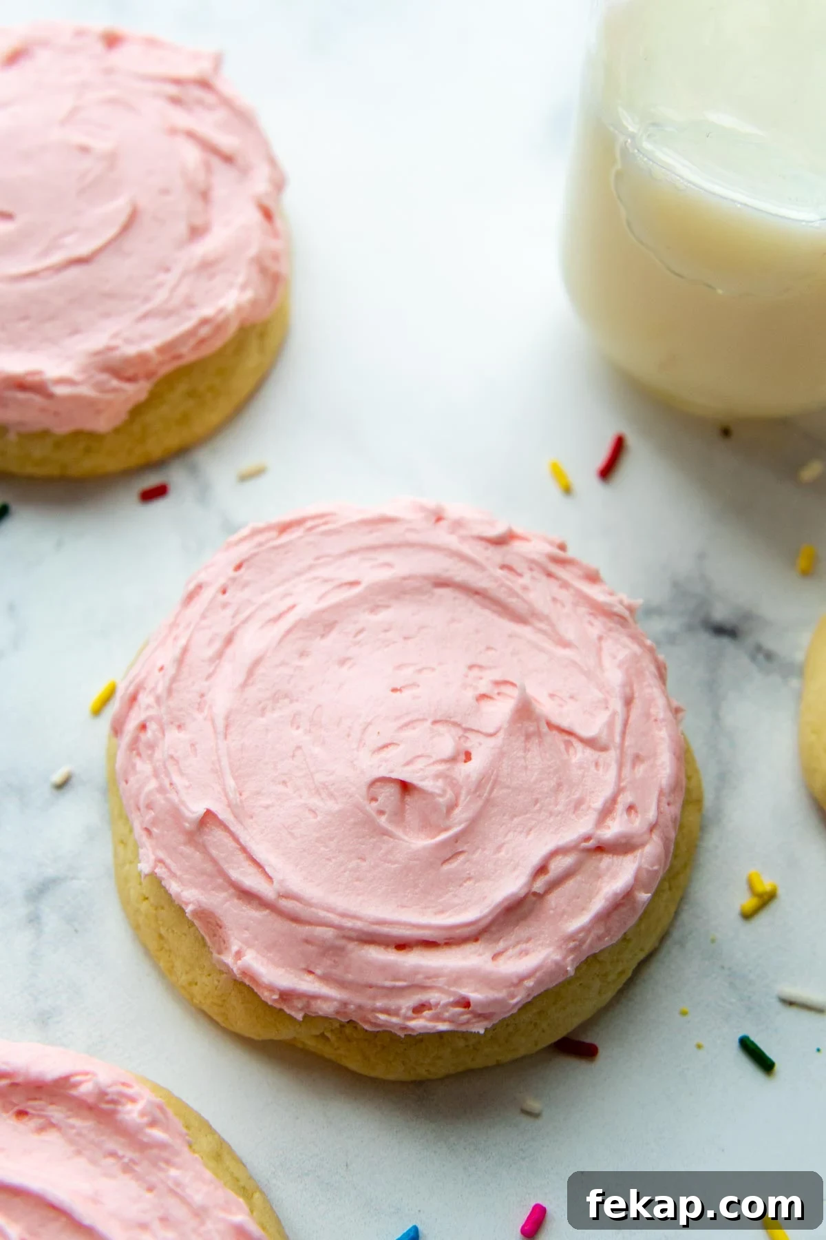 a pink sugar cookie on a white background with sprinkles around and a glass of milk.