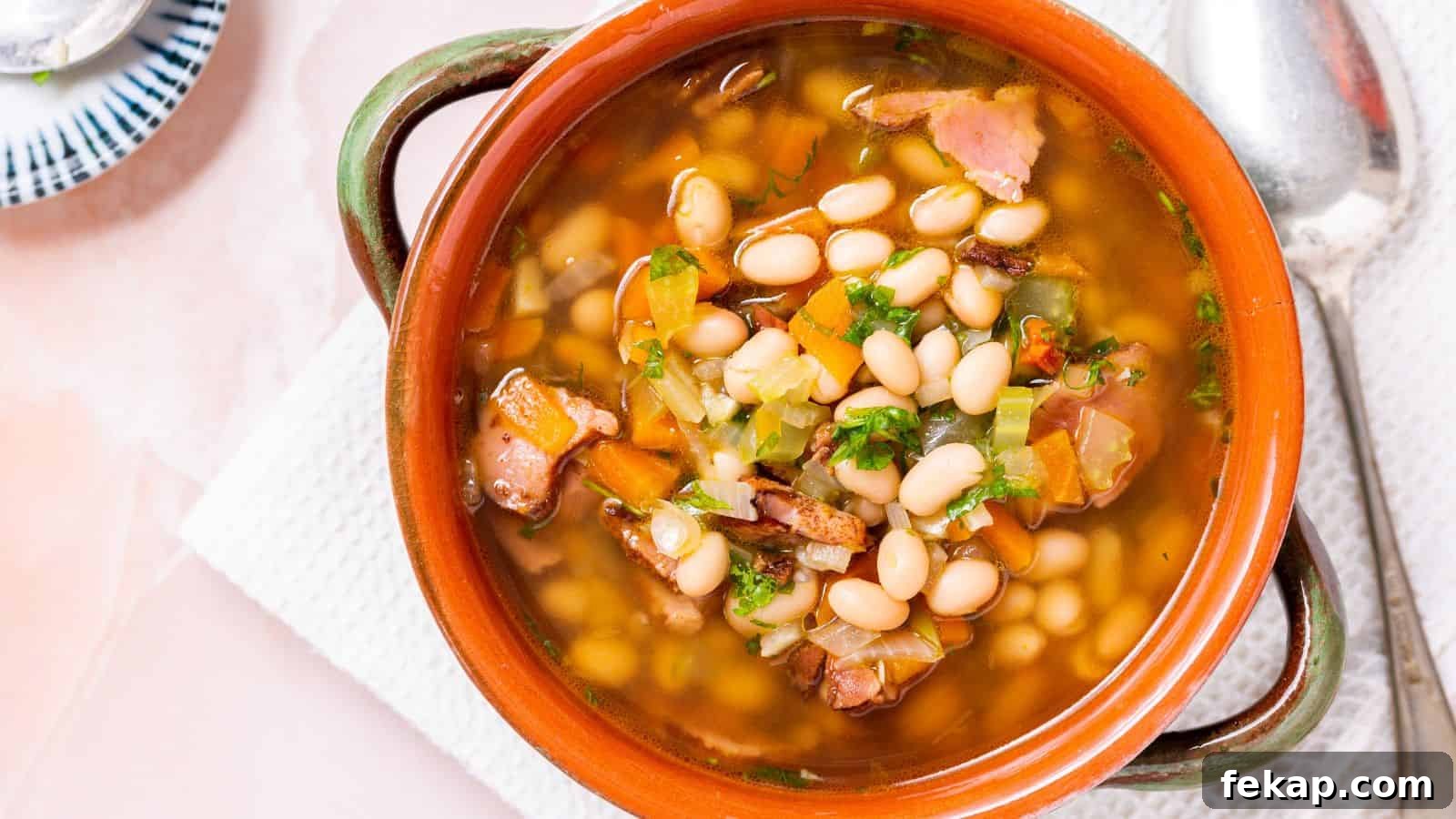 15 Soups and Stews That Make You Forget It's Summer 9 A bowl of bean soup with chunks of ham and diced vegetables, served in a rustic terracotta bowl with a spoon on the side.