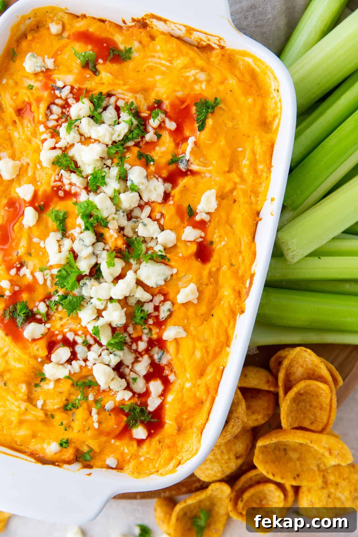 Buffalo Chicken Dip garnished with blue cheese and Frank's Red Hot in a baking dish next to celery and Frito's Scoops.