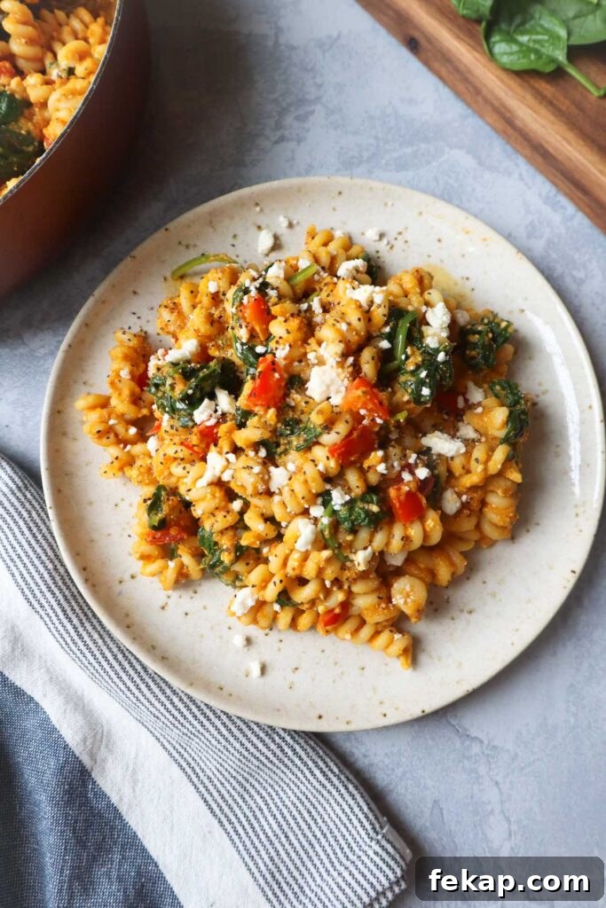 white plate with pasta tossed in a pumpkin feta sauce topped with red bell pepper and spinach