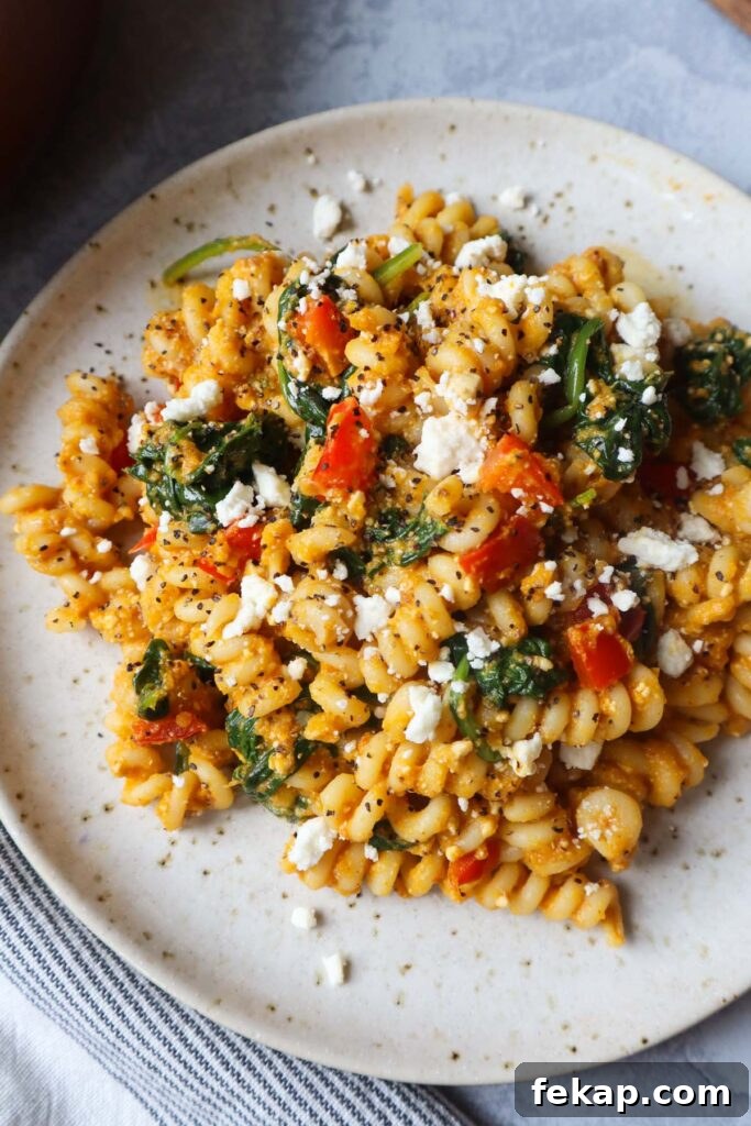 white plate with pasta tossed in a pumpkin feta sauce topped with red bell pepper and spinach