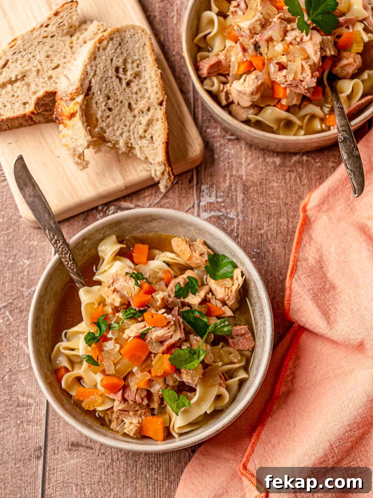 Two bowls of turkey noodle soup with egg noodles, bread slices, and a peach napkin.