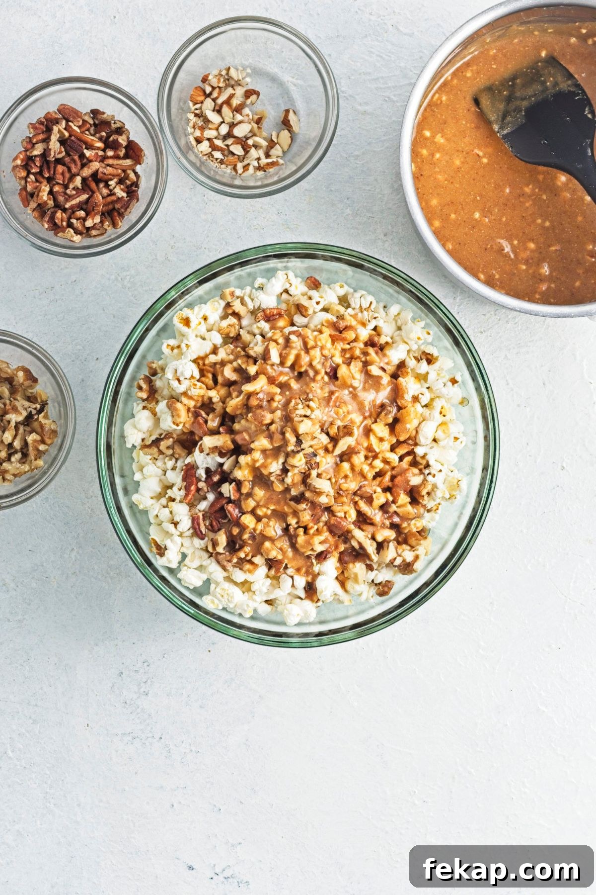 Caramel sauce and chopped nuts on top of popcorn in a glass bowl with ingredients in bowls around it.