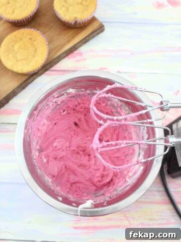 Pink cream cheese frosting in a steel bowl with a handheld blender over top and cupcakes on a cutting board behind them.