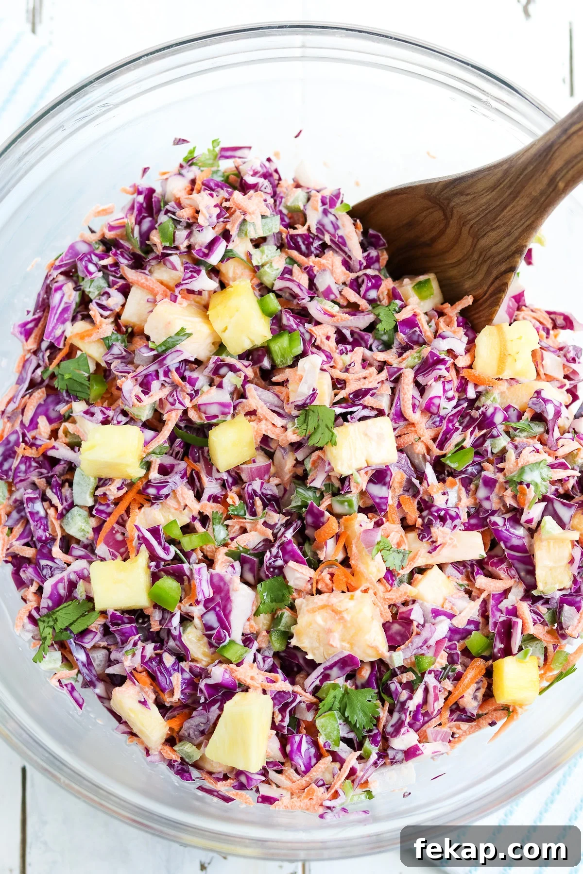 wooden spoon tossing together colorful pineapple jalapeno coleslaw in glass bowl.