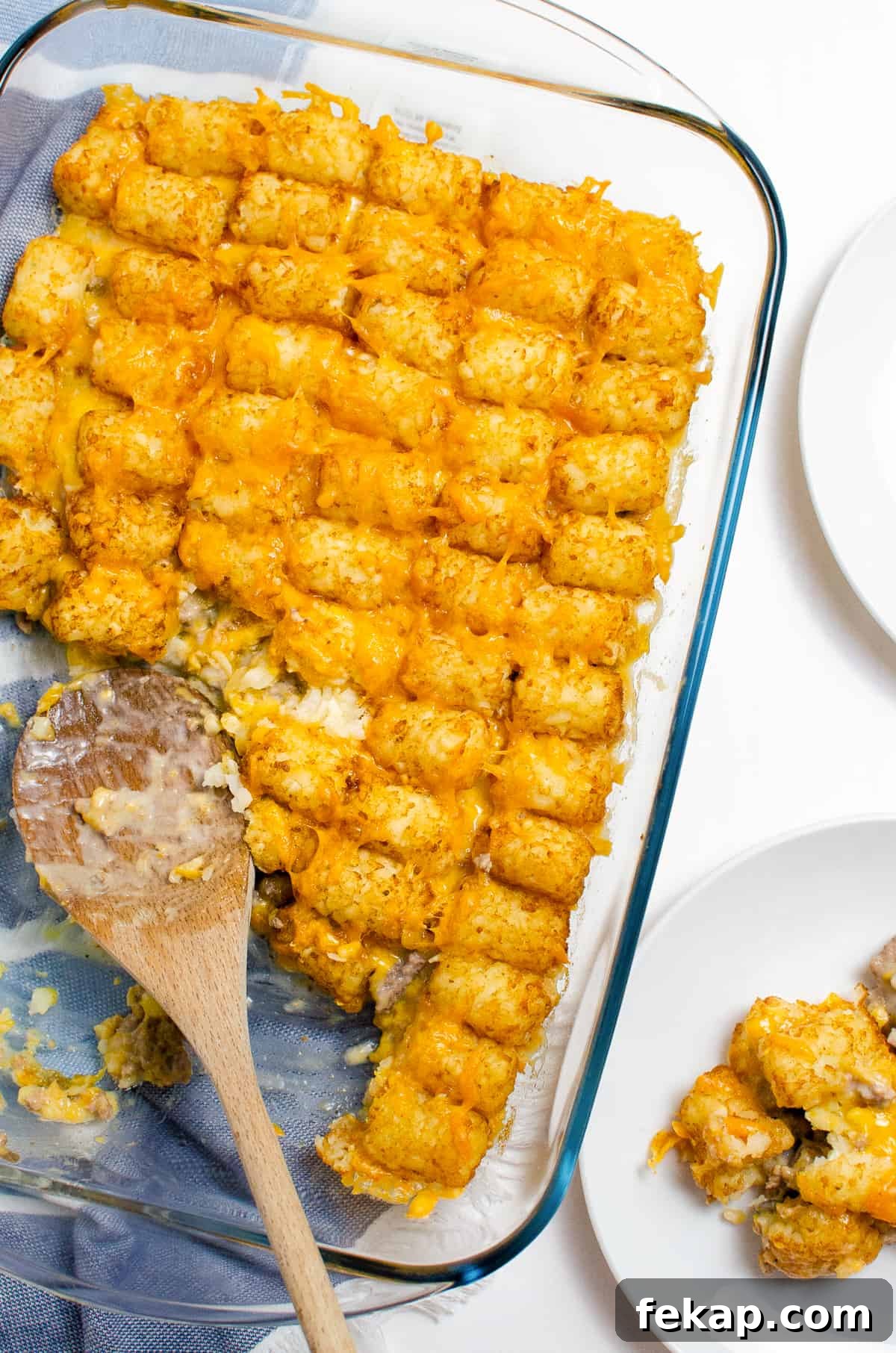 Tater tot casserole with ground beef and melted cheese in a 9 x 13 dish. A wooden spoon in resting in the half-empty dish and a serving of the casserole can be seen on a white plate halfway out of the frame