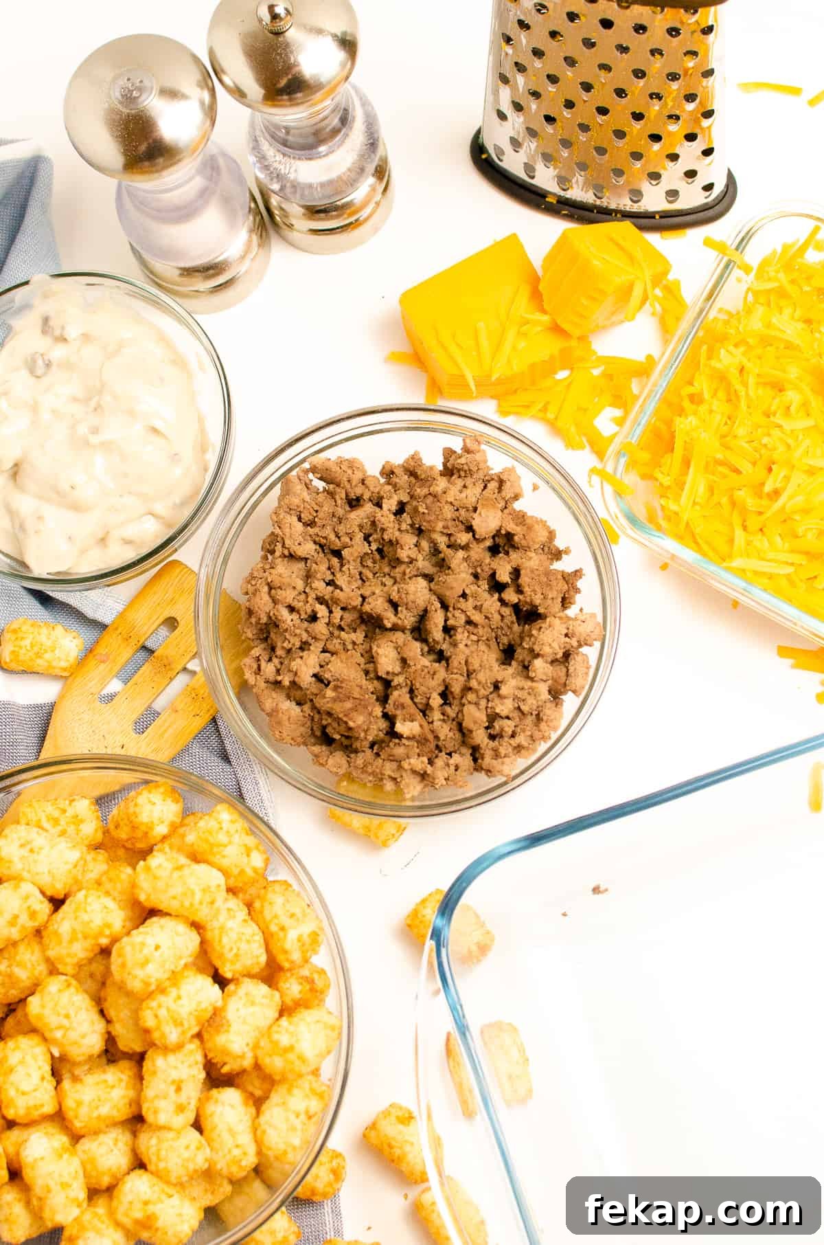 Ingredients in glass bowls: browned ground beef, cream of mushroom soup, cheddar cheese and cheese shredder, frozen tater tots, salt and pepper, and glass casserole dish