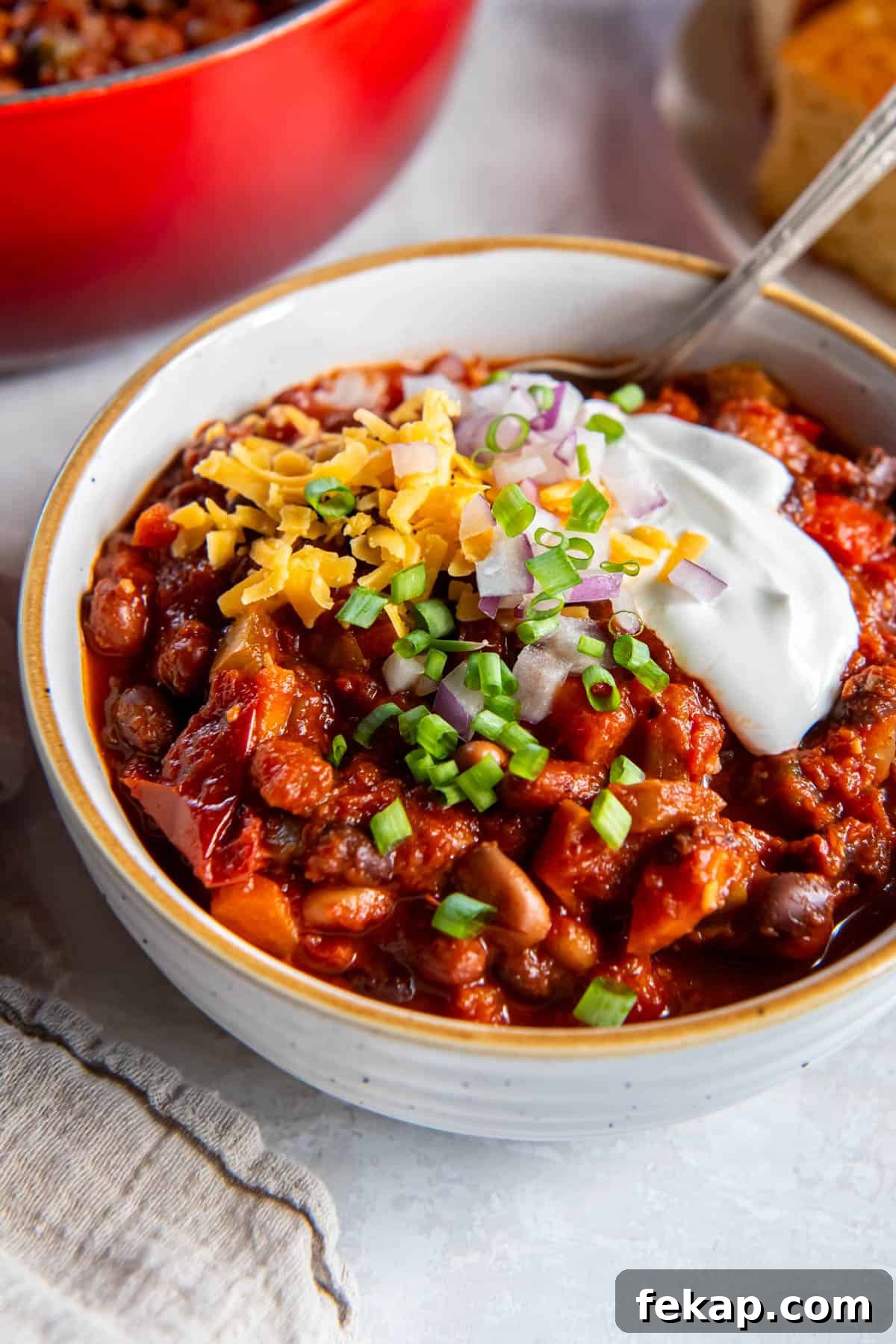 A bowl filled with three bean chili topped with sour cream, cheddar cheese, red onion and chives, with a spoon for serving.