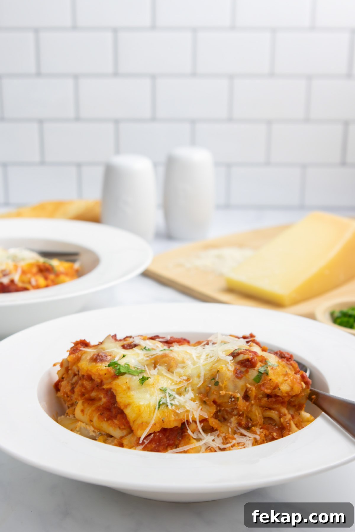 A serving of cheesy meatless baked ziti in a bowl with a fork with a cutting board with parmesan cheese in the background.