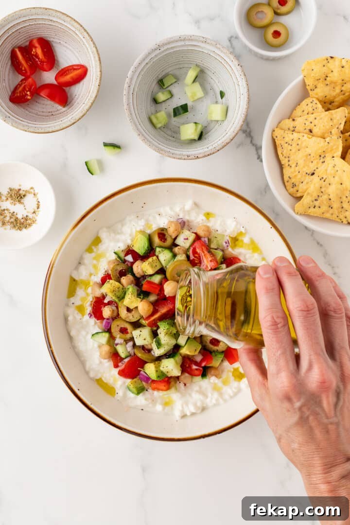 Woman's hand pouring olive oil over a cottage cheese greek salad in a white bowl.