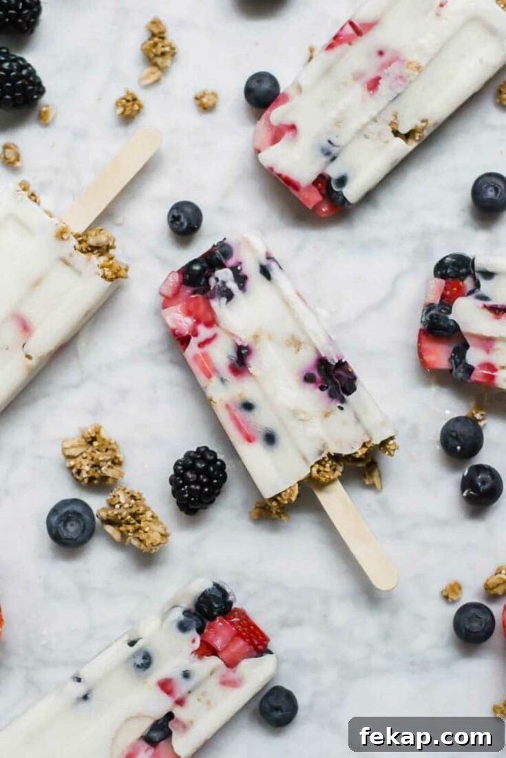 Several popsicles laying at various angles on marble counter, with berries and granola scattered in between.