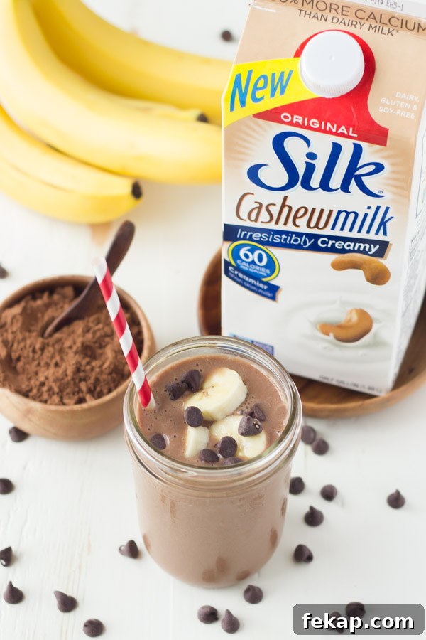 Chocolate Banana Smoothie topped with banana slices and chocolate chips, in a glass jar with a straw, sitting next to a small bowl of cocoa powder, a bunch of whole bananas and a carton of Silk Cashewmilk.