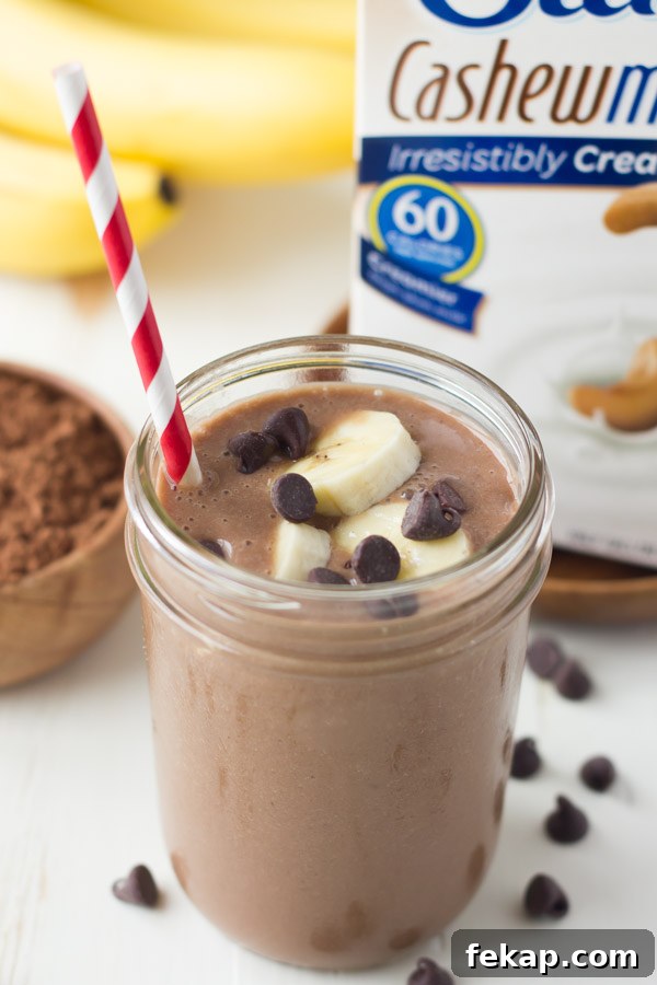 Chocolate Banana Smoothie topped with banana slices and chocolate chips, in a glass jar with a straw, with a small bowl of cocoa powder, a bunch of whole bananas and a carton of Silk Cashewmilk in the background.