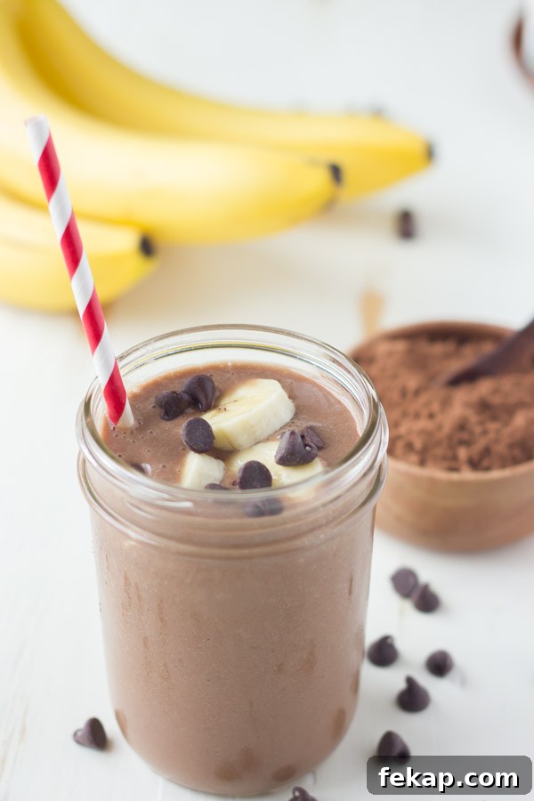 Chocolate Banana Smoothie topped with banana slices and chocolate chips, in a glass jar with a straw, with a small bowl of cocoa powder and a bunch of whole bananas in the background.