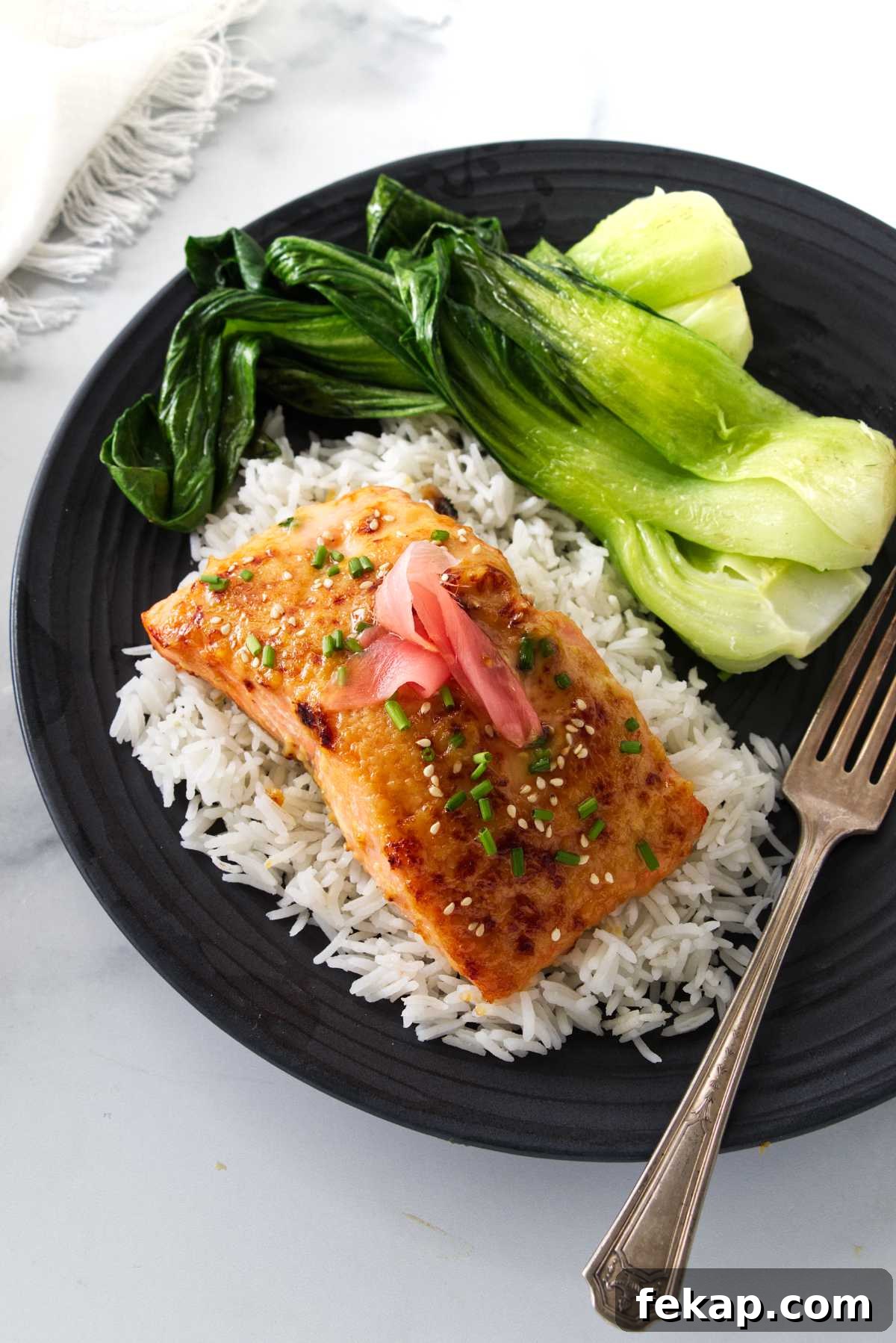 Overhead view of a plated serving of miso butter salmon on a mound of rice and steamed bok choy. A dinner fork on the plate and a napkin to the side.