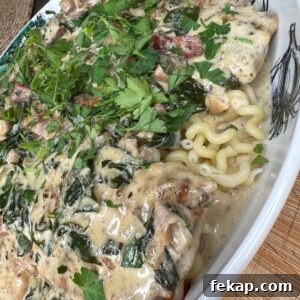 Creamy Marry Me Chicken Recipe 4 A close-up of creamy pasta topped with chopped herbs, spinach, diced tomatoes, and a rich white sauce, served in a white dish. The pasta is curly and the sauce covers the ingredients generously.