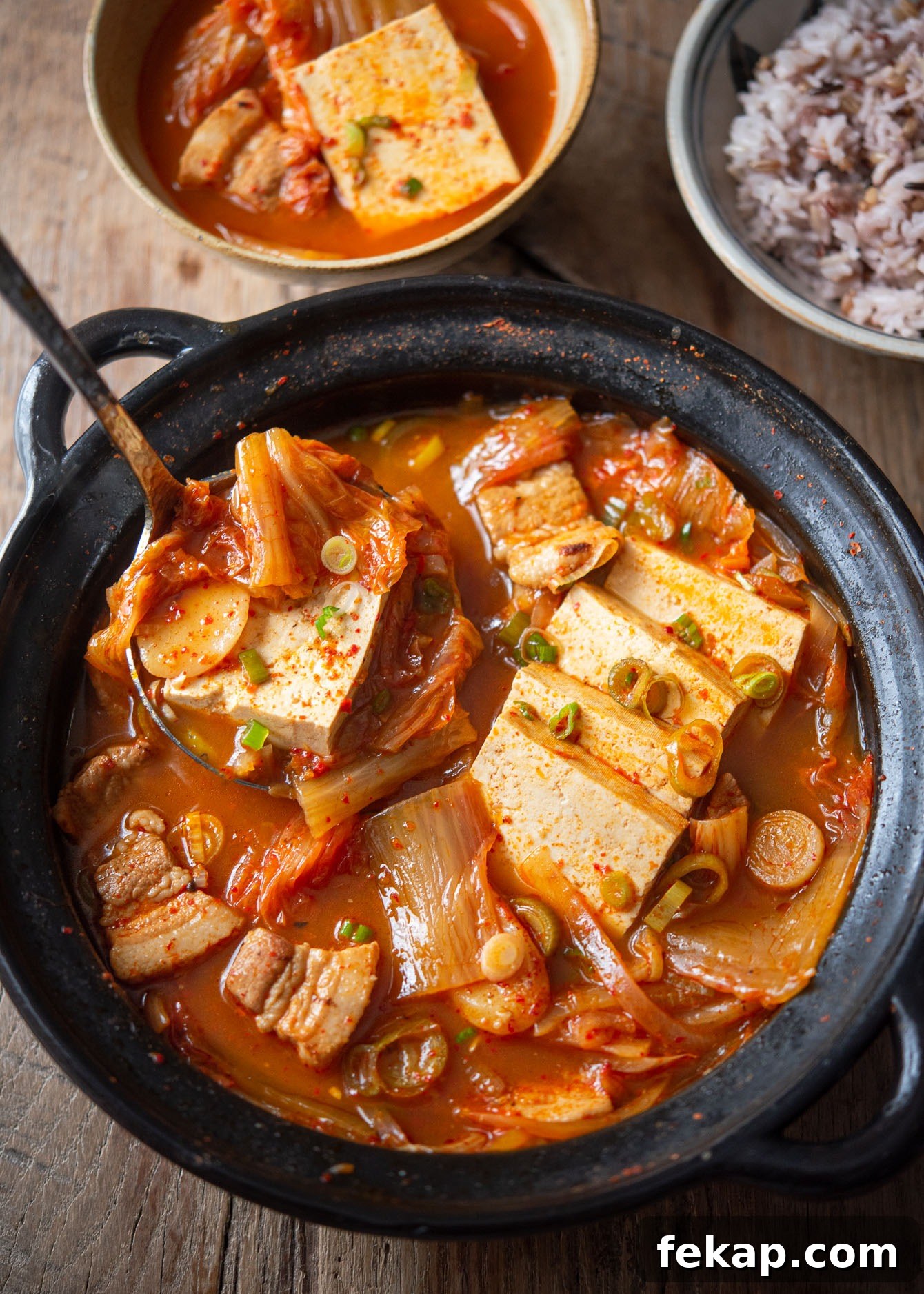 Ladle lifting kimchi jjigae with tofu and pork belly from a simmering pot.