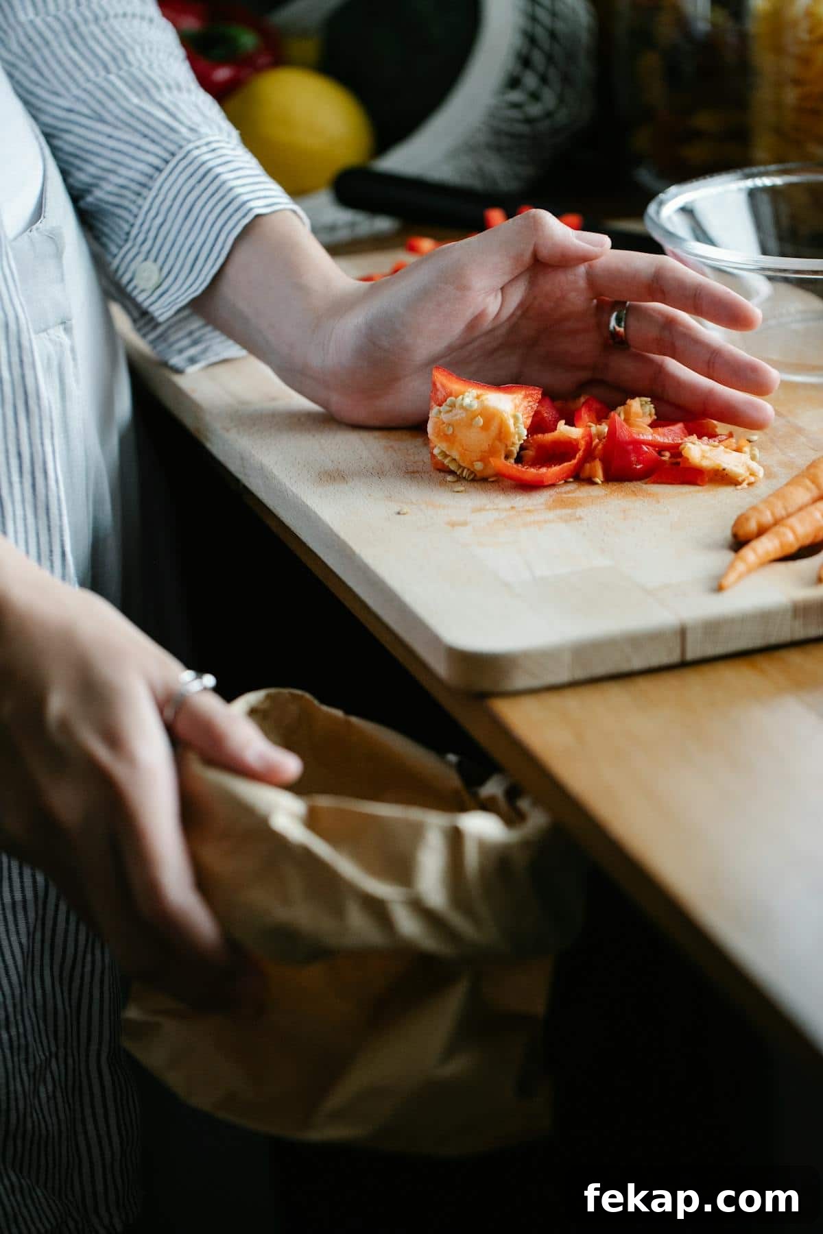 Broth Stock and Bone Broth Differences Explained 3 A person preparing food on a wooden kitchen counter, with sliced red peppers and an orange on a cutting board. They are planning to make homemade vegetable broth and are holding a brown paper bag for scraps, wearing a striped shirt. Carrots are also visible nearby.