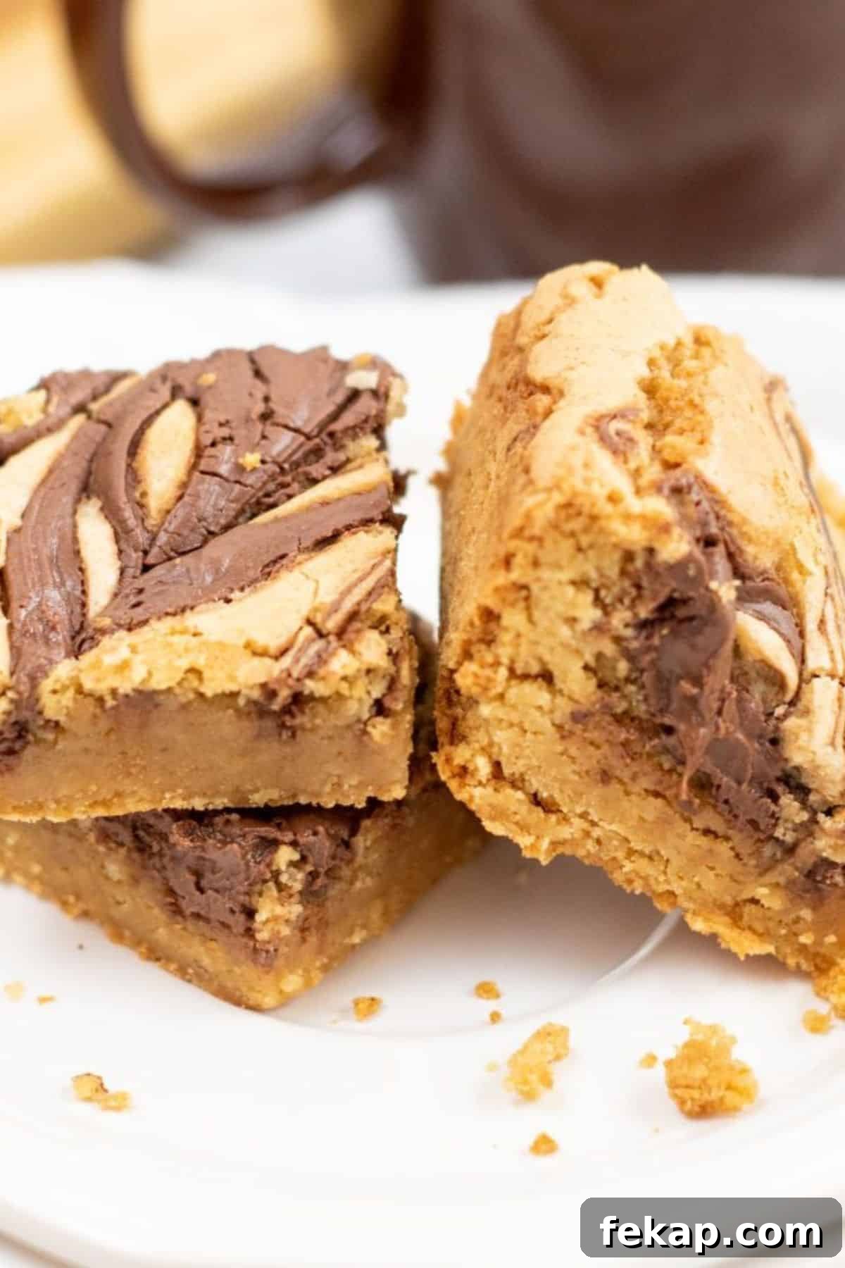 Two thick Brown Butter Blondies with a chocolate swirl on top are stacked on a white plate, with crumbs scattered around them.