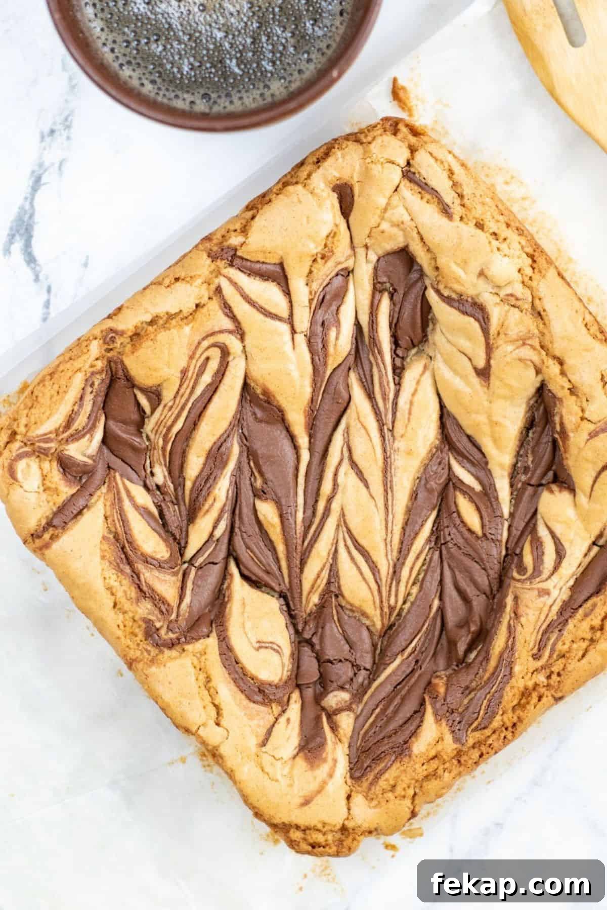 A tray of swirled chocolate and brown butter blondies sits on parchment paper beside a cup of black coffee on a marble surface.