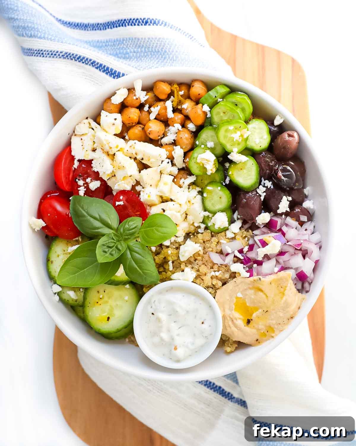 Greek Quinoa Bowl Recipe with Feta, Cucumber and Olives 2 A white bowl filled with quinoa, fresh vegetables, basil, feta cheese, chickpeas, and tzatziki.