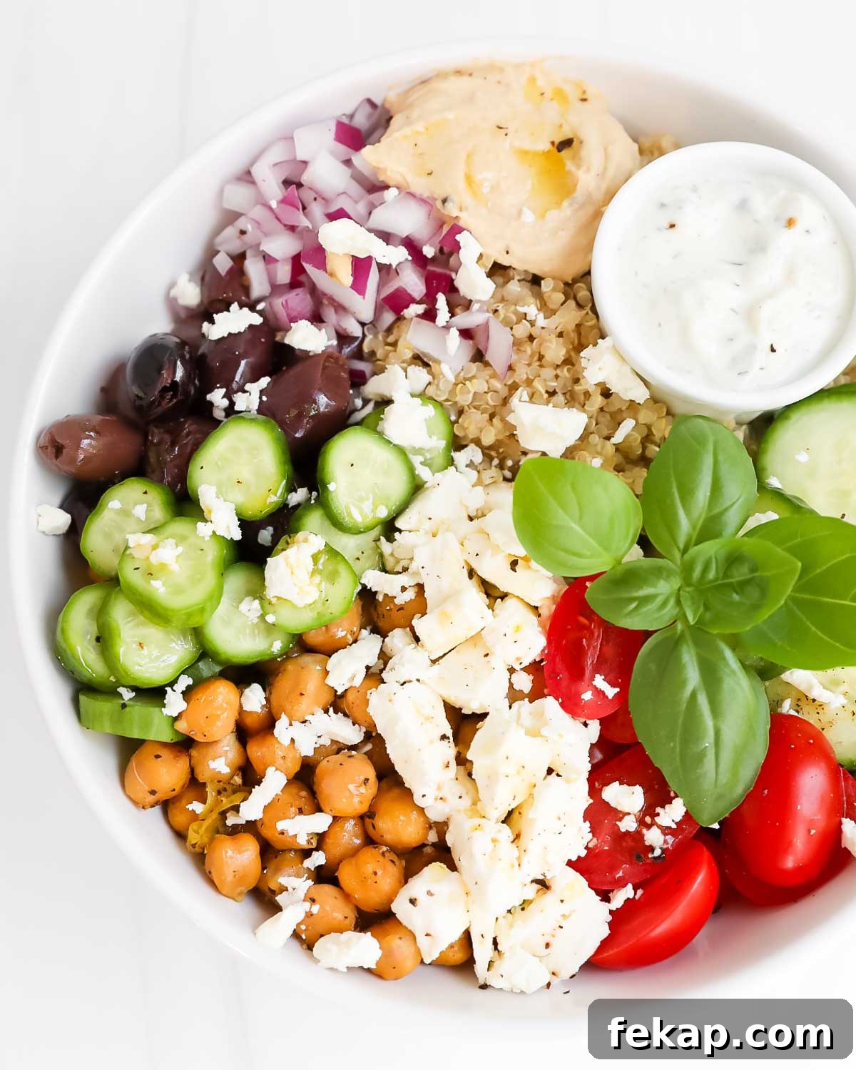 Greek Quinoa Bowl Recipe with Feta, Cucumber and Olives 5 A bowl filled with quinoa, vegetables, chickpeas, hummus, tzatziki, fresh basil, and feta cheese.