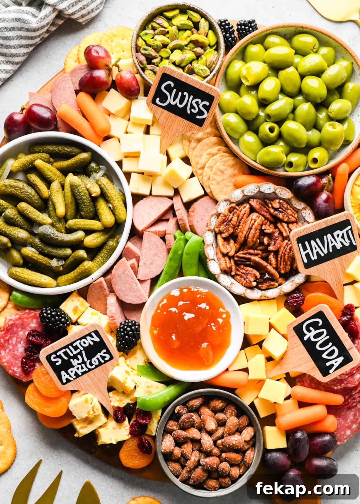 up close overhead photo of a charcuterie board with assortment of cubed cheeses with chalkboard labels reading Swiss, Gouda, Havarti and Stilton with apricots), green olives, pub cheese spread, salami, summer sausage, pickles, nuts, dried and fresh fruits, crackers, and a bowl of apricot jam, all arranged on a round wooden serving board.