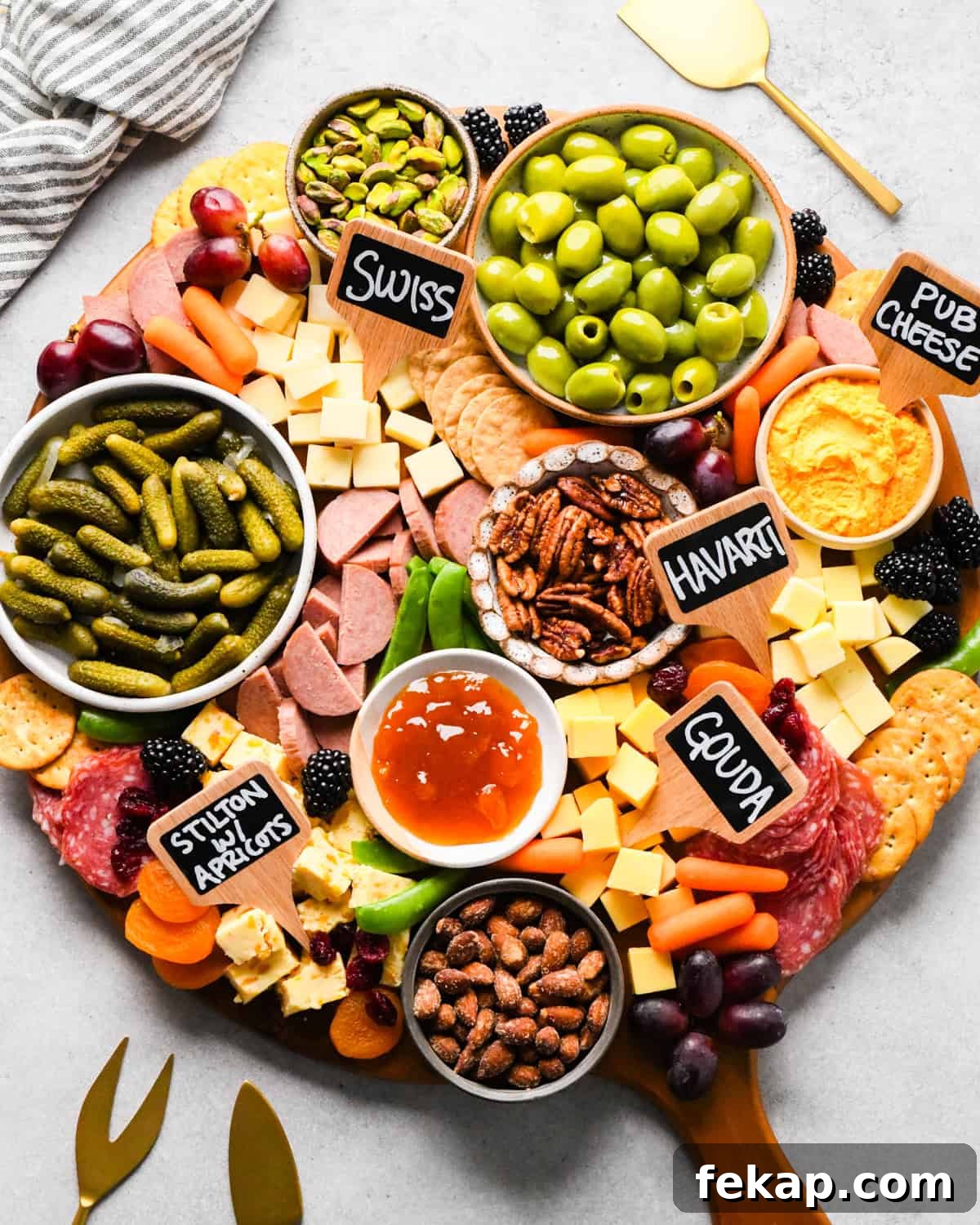 overhead photo of a charcuterie board with assortment of cubed cheeses with chalkboard labels reading Swiss, Gouda, Havarti and Stilton with apricots), green olives, pub cheese spread, salami, summer sausage, pickles, nuts, dried and fresh fruits, crackers, and a bowl of apricot jam, all arranged on a round wooden serving board.