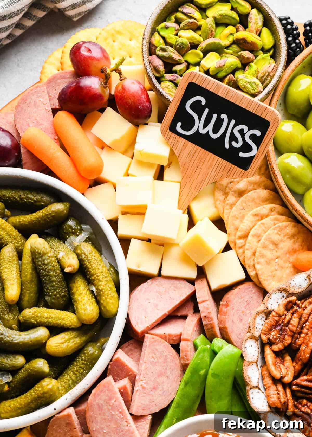 up close photo of a portion of a charcuterie board with a cheese marker labeled "swiss," pickles, pistachios, grapes, carrots, summer sausage, green olives and pecans