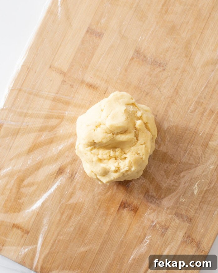Cut Out Sugar Cookie Recipe for Perfect Shapes 14 Sugar cookie dough rolled into a ball on top of plastic wrap on a cutting board.