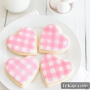 Cut Out Sugar Cookie Recipe for Perfect Shapes 10 A plate of four heart-shaped cookies decorated with pink and white gingham icing. The plate is on a white cloth. In the background, there are eggs, a glass of milk, and a jar. The setting is on a white wooden surface.